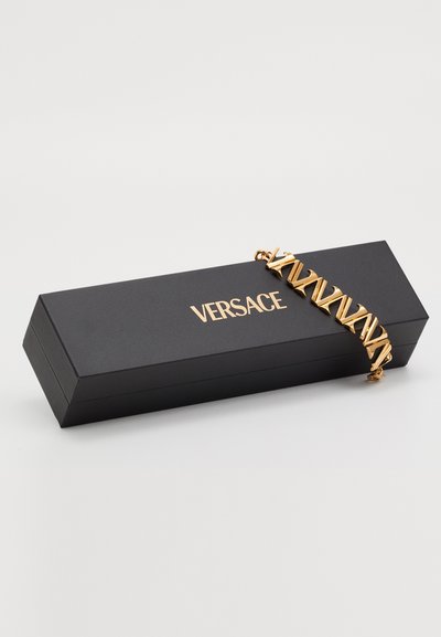 Kuldmetallist käevõru, millel on omavahel seotud geomeetriline disain, esitatud mustal ristkülikulisel Versace karbil, millel on kuldne logo.