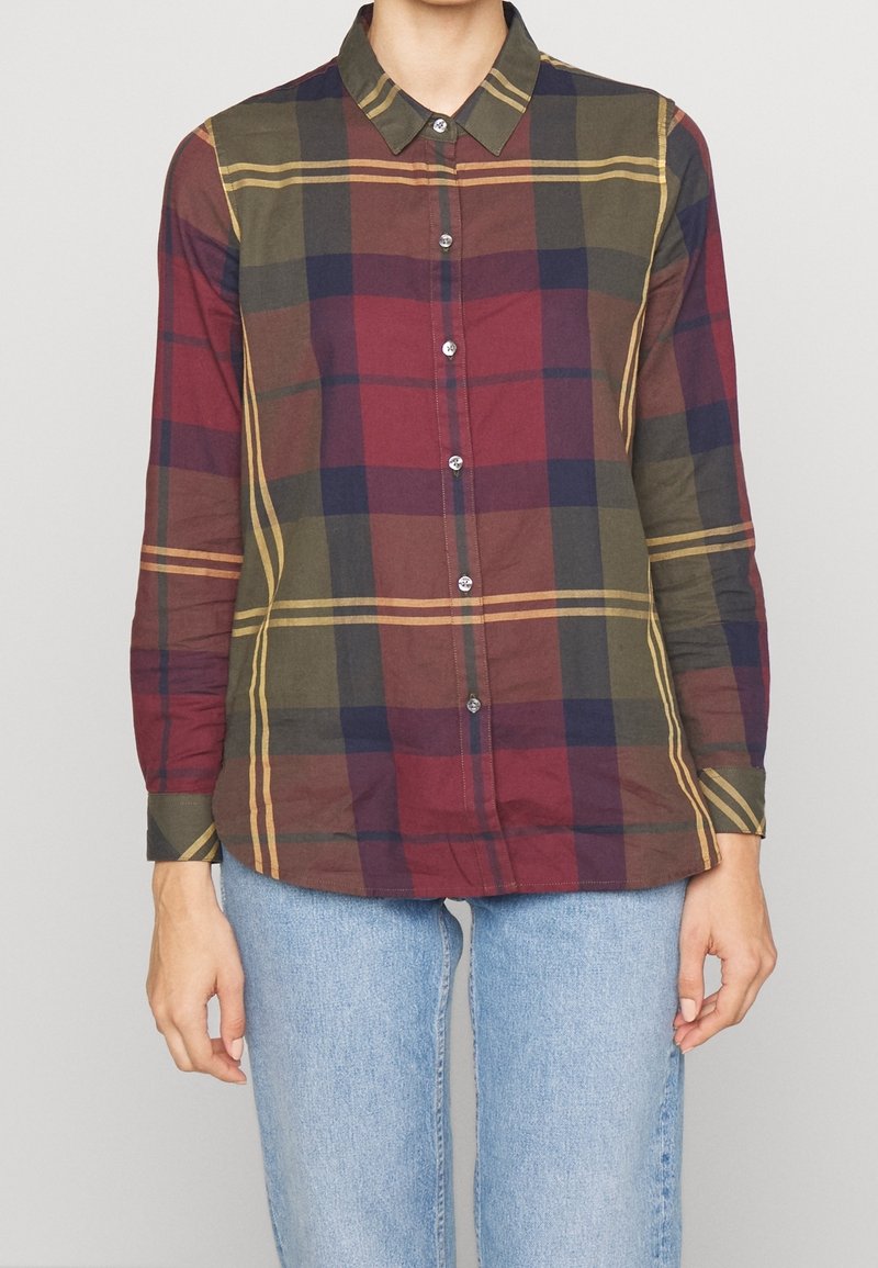 Personne portant une chemise à manches longues boutonnée avec un motif à carreaux vert, rouge, marine et jaune, et un jean bleu clair, debout devant un fond uni.