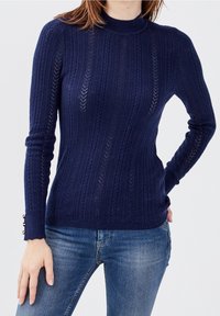 Navy modrý pletený svetr s vertikálním žebrováním, vysokým límcem a vlnitými manžetami, na kterých se nacházejí dekorativní knoflíky.