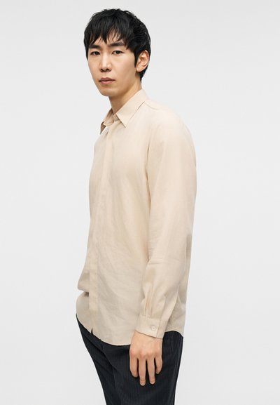 Μπεζ πουκάμισο μακρυμάνικο με κλασικό γιακά, απαλό ύφασμα και μανσέτες με κουμπιά, σε συνδυασμό με σκούρο παντελόνι με λεπτές ρίγες.