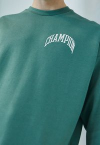 Zelená bavlněná mikina s prohnutým bílým logem "CHAMPION" na přední straně. Klasický kulatý výstřih a uvolněný střih, s texturovanou látkou.