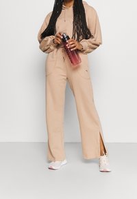 Persona che indossa un felpa beige e pantaloni larghi, tiene in mano una bottiglia d'acqua rosa, in piedi su uno sfondo bianco semplice.