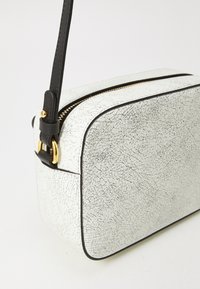 Borsa a tracolla in pelle bianca testurizzata con zip color oro, tracolla nera e forma rettangolare. Presenta un motivo superficiale screpolato.