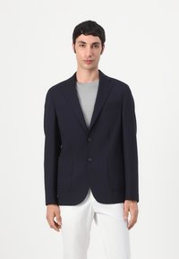 Tengerészkék blézer két elülső zsebbel, egygombos záródással, kihajtott gallérral és sima felülettel. Szürke póló és fehér nadrág fölött viselve.