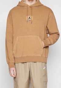 Felpa con cappuccio marrone dotata di tasca a marsupio, caratterizzata da un piccolo patch con logo e coulisse. Il tessuto sembra morbido con una finitura testurizzata.