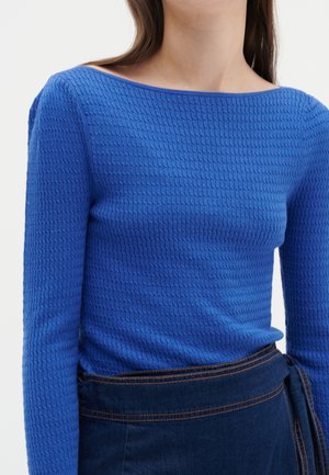 Haut long bleu à texture avec un large col. Dispose d'un motif côtelé et d'une silhouette ajustée, associé à une jupe en denim foncé.