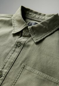 Close-up van een khaki groene blouse met kraag en een voorste borstzak, waarop de fijne stiksel en knopen duidelijk zichtbaar zijn.