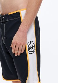 Černé plavkové šortky s bílými a žlutými akcenty, zdobené logem a kontrastním prošíváním. Materiál působí lehce a rychleschnoucí.