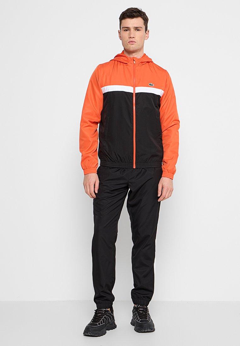 Coupe-vent multicolore avec une partie supérieure orange, une partie inférieure noire et une bande horizontale blanche. Il dispose d'une capuche, d'une fermeture éclair et de poignets élastiques.