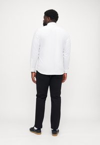 Camicia bianca a maniche lunghe con orlo curvo e polsini con bottoni, abbinata a pantaloni neri e sneakers blu navy, vista da dietro.
