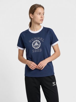 Kvinde iført mørkeblå Hummel sportst-shirt med hvide kanter og sorte Hummel bukser, ser mod venstre med hænderne foldet.