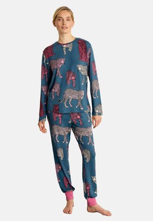 Femme portant un haut à manches longues bleu sarcelle et un pantalon à imprimé léopard rose et blanc avec des poignets roses aux chevilles, debout pieds nus sur fond blanc.