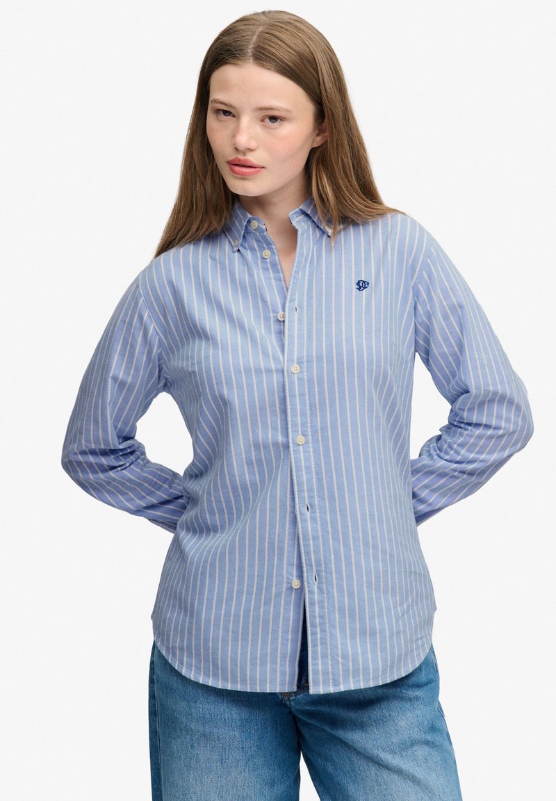 Giovane donna con capelli lunghi che indossa una camicia a righe blu e bianca con bottoni e jeans blu, in piedi con le mani dietro la schiena.