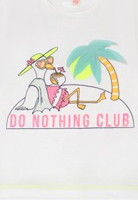 A camisola branca apresenta uma ilustração de um pássaro relaxado com óculos de sol, uma palmeira e "CLUBE DO NADA" em letras brilhantes rosas.