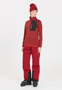 Rote Outdoor-Jacke mit schwarzem Brusttasche und orangefarbenem Reißverschluss, kombiniert mit roten Hosen und schwarzen Stiefeln. Verfügt über einen hohen Kragen und strukturiertes Gewebe.