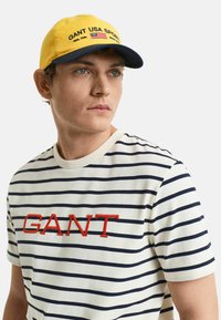Gorra amarilla con visera azul marino, bordado "GANT USA SPORTS," llevada con una camiseta de rayas azul marino y blanca que presenta el texto rojo "GANT".