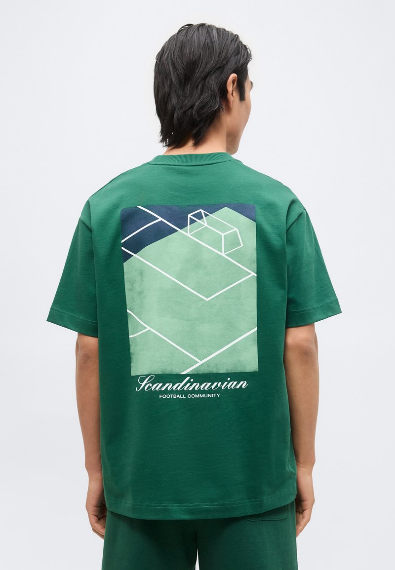 Άνδρας φοράει πράσινο t-shirt με εικόνα γηπέδου ποδοσφαίρου και το κείμενο "Κοινοτητα σκανδιναβικού ποδοσφαίρου" στο πίσω μέρος.