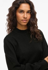 Νεαρή γυναίκα με κυματιστά σκούρα μαλλιά, φορώντας ένα μαύρο πλεκτό πουλόβερ, που κοιτάζει ήρεμα την κάμερα μπροστά σε ένα απλό λευκό φόντο.