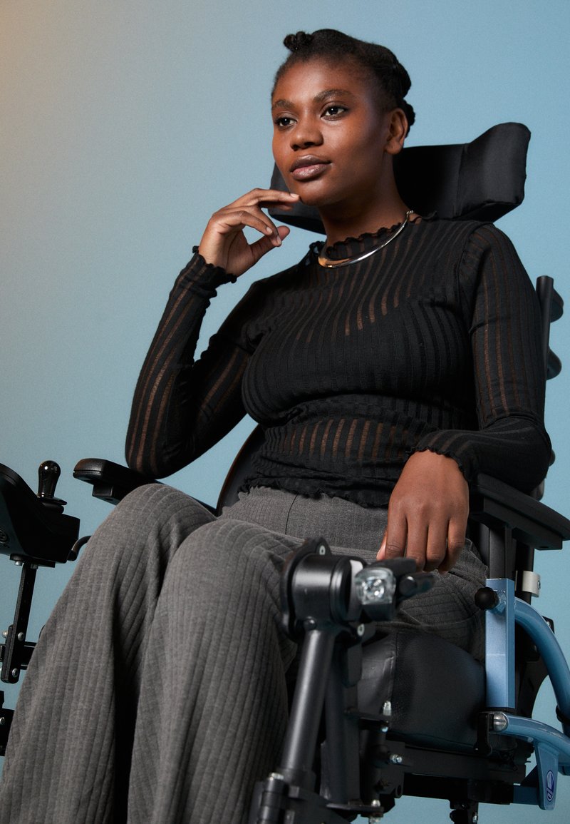 Top preto de mangas longas com riscas verticais transparentes, combinado com calças largas cinzentas de malha canelada. Sujeito sentado numa cadeira de rodas moderna com características ajustáveis.