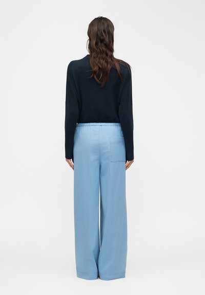 Femme aux longs cheveux bruns, portant un haut noir à manches longues et un pantalon ample bleu clair, debout de dos sur un fond uni.