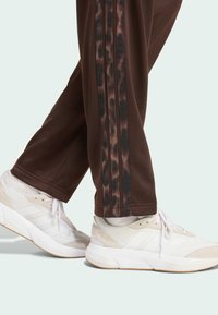 Pantaloni sportivi marroni con strisce laterali nere e a stampa leopardata, abbinati a sneakers bianche con dettagli in suede grigio e suole testurizzate.