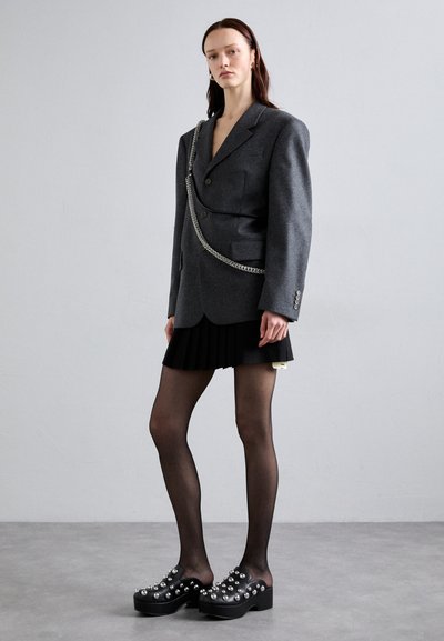 Gray wool blazer with structured shoulders, paired with a black pleated skirt and black clogs adorned with metallic studs. Black tights.