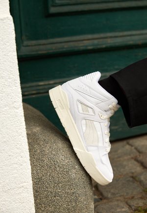 Zapatillas blancas de caña alta con cuero texturizado, acentuadas con perforaciones sutiles y una suela crema, descansando sobre una superficie lisa de piedra.