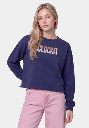 Młoda kobieta z długimi blond włosami nosi granatową bluzę z tekstem "CLOCK IT" oraz jasnoróżowe spodnie, pozując na jednolitym tle.