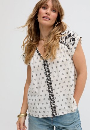 Frau mit welligem blondem Haar, die eine weiße kurzärmelige Bluse mit Muster und blaue Jeans trägt, dazu ein goldenes Armband und eine Halskette, schaut leicht nach oben.