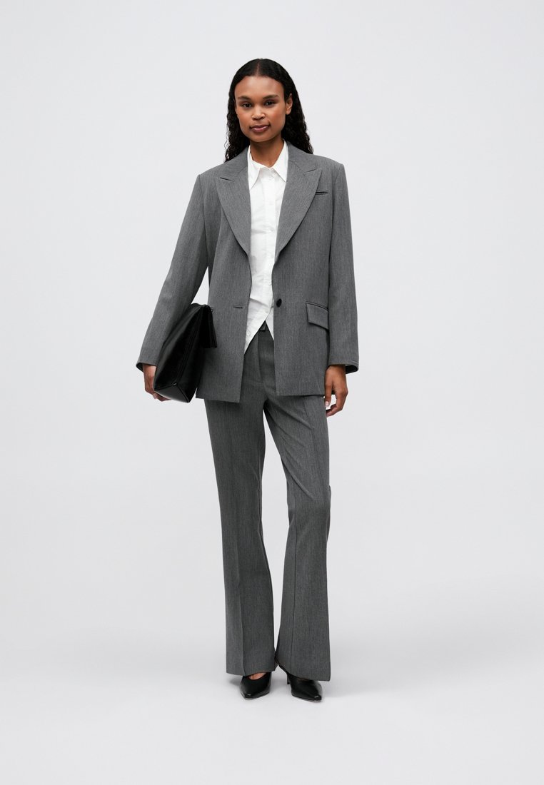Costume gris ajusté avec un blazer à bouton unique, un pantalon à jambes larges, une chemise blanche en dessous et une pochette noire. Texture de tissu lisse.
