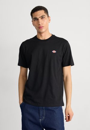 Jeune homme portant un t-shirt noir à manches courtes avec un petit logo Dickies sur la poitrine et un jean bleu, debout devant un fond uni.