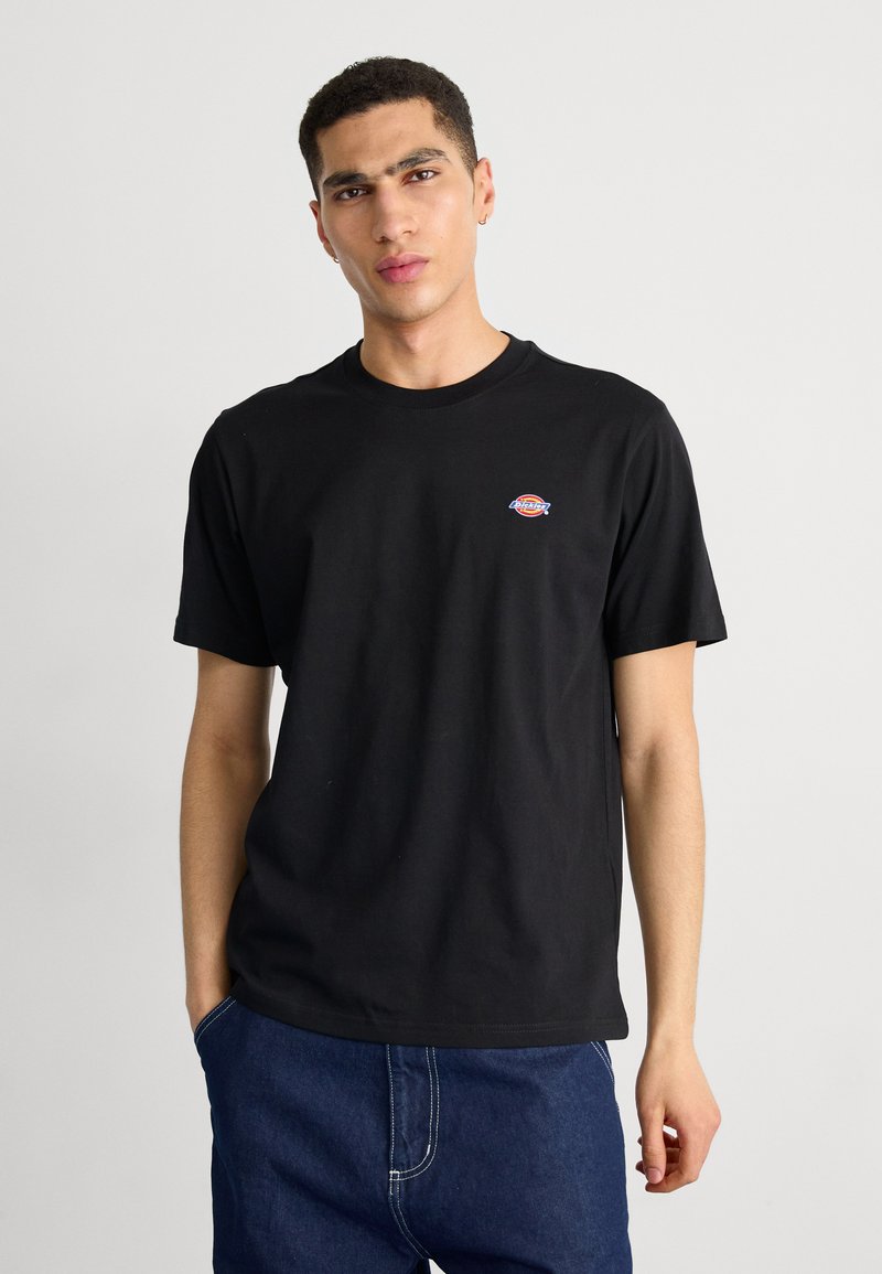 Ung mand iført en sort kortærmet t-shirt med et lille Dickies-logo på brystet og blå jeans, stående mod en ensfarvet baggrund.