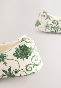 Pantoffeln mit Blumenmuster aus weichem cremefarbenem und grünem Stoff, mit geschwungenem Design und einem dekorativen Etikett an einer Seite.