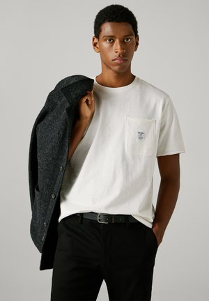 Camiseta blanca de algodón con un bolsillo en el pecho y etiqueta, combinada con pantalones negros y un abrigo de lana gris oscuro sostenido sobre el hombro.