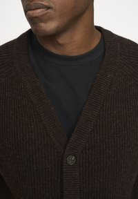 Tummanruskea ribbetty cardigan V-pääntie-mallilla, jossa on yksi napitus. Päällä musta pyöreä pääntie paita.