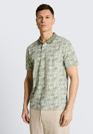 Homme portant un polo à manches courtes vert et blanc à motif de palmiers et un pantalon beige, debout devant un fond uni.