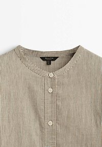 Chemise beige et noire à rayures verticales, boutonnée, avec col rond, trois boutons visibles et une étiquette en tissu à l'intérieur de l'encolure.