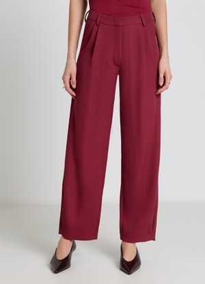 Donna con pantaloni bordeaux a vita alta e gamba larga abbinati a tacchi neri a punta, in piedi su uno sfondo chiaro e uniforme.