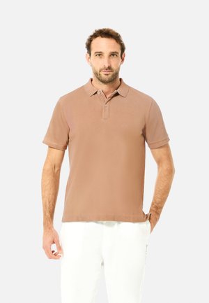 Homme portant un polo à manches courtes marron clair et un pantalon blanc, debout devant un fond clair uni.