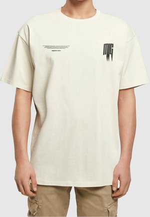 Mand iført en lys beige oversized T-shirt med lille sort tekst på venstre bryst og sort stiliseret logo på højre bryst, kombineret med beige cargobukser.