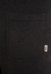 Czarna koszulka z materiału z kieszonką na klatce piersiowej, wzmocnionymi szwami i małą białą naszywką Vans w dolnej części. Miękka faktura.