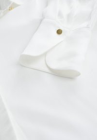 Biela blúzka z hladkej látky s ostrým golierom a jedným zlatým gombíkom na manžete. Mäkka textúra a čisté línie.