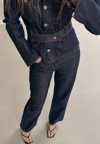 Tamsiai mėlynos denim striukės ir kelnių rinkinys su raudonais siūlais, pasižymintis dirželiu ant juosmens, didelėmis priekinėmis kišenėmis ir sidabro mygtukų akcentais.