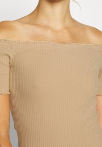 Haut beige clair épaules dénudées avec texture côtelée, manches courtes et bords effilochés. Confectionné en tissu extensible, design ajusté.