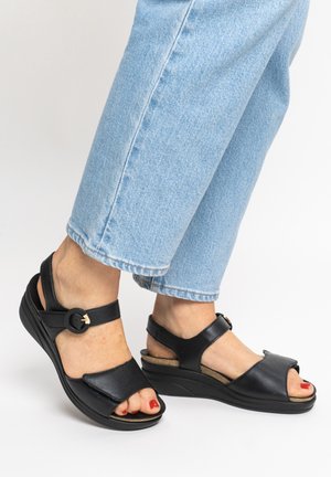Feet wearing black open-toe wedge sandals with ankle straps and light blue jeans, standing on a white surface.