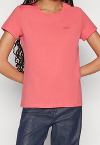 Kvinna klädd i korallrosa t-shirt med kort ärm och liten broderad "BOSS"-logotyp på bröstet samt mörkblå byxor, med händerna bakom ryggen.