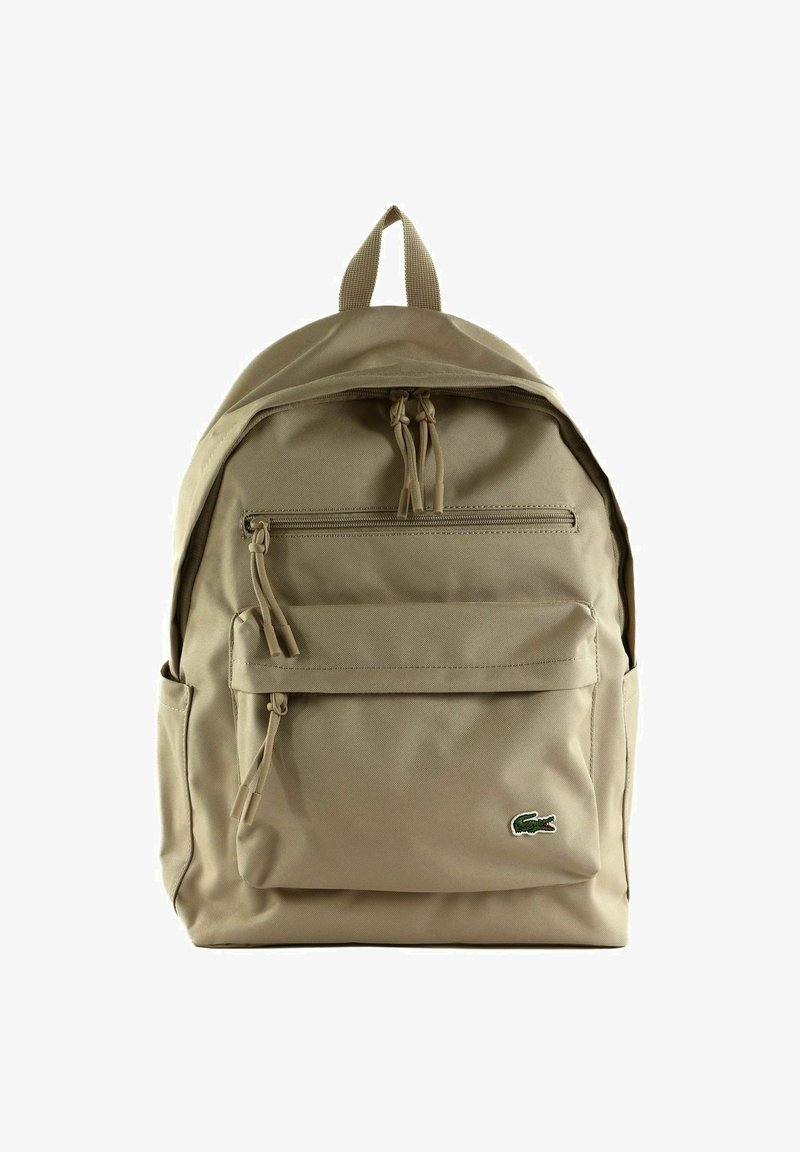 Sac à dos beige avec deux compartiments zippés, poches latérales, poignée supérieure et petit logo vert de crocodile sur la poche avant.