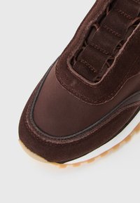 Gros plan sur l'avant d'une sneaker marron avec des superpositions en suède, un dessus en tissu, des coutures visibles et une semelle en caoutchouc beige sur fond blanc.