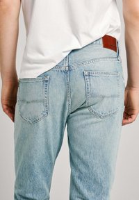 Jeans de denim azul claro con un ajuste relajado, que cuentan con dos bolsillos traseros y un parche de cuero marrón en la cintura. Textura suave y desgastada.