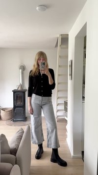 Svart cardigan med perleknapper, grå vide bukser og svarte ankelstøvler. Innendørs setting med tregulv og enkel dekor.